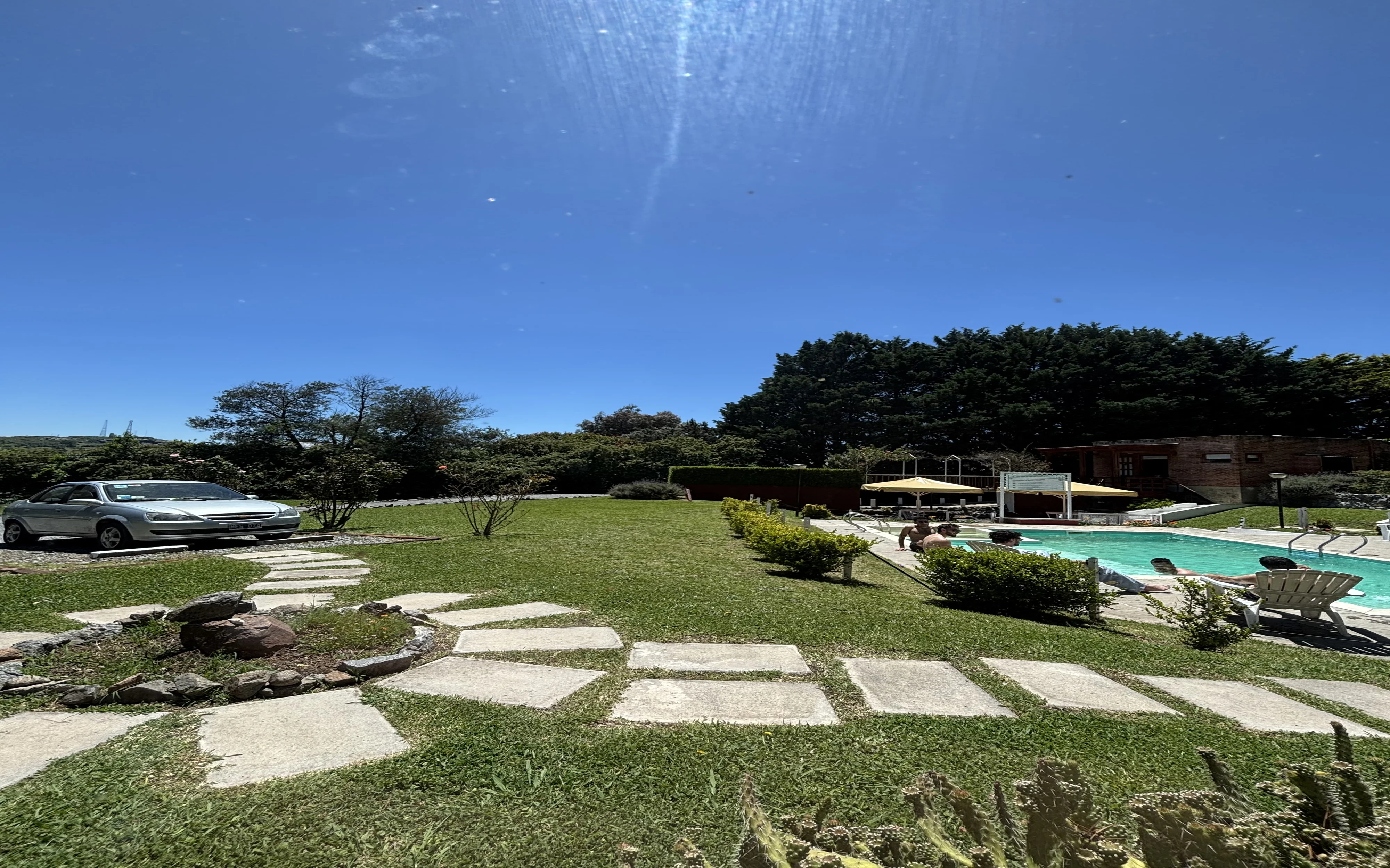 pileta con gente en solarium y auto estacionado